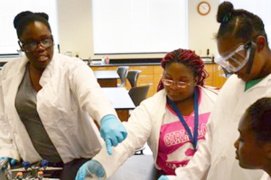 Girls For A Change After School Program Woman working in lab coat helping girls with safety glasses on