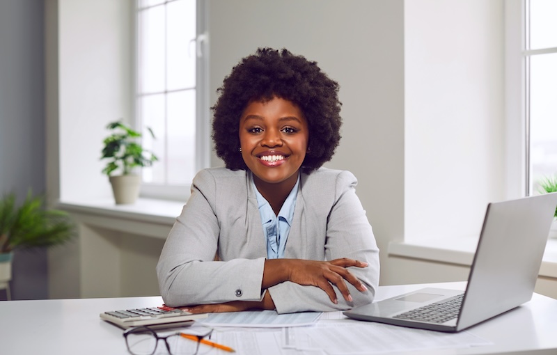 Portrait of happy young African American businesswoman or financial accountant at office desk Portrait of happy young businesswoman or financial accountant. Beautiful African American woman looking at camera and smiling while sitting at her desk with laptop computer in her business office